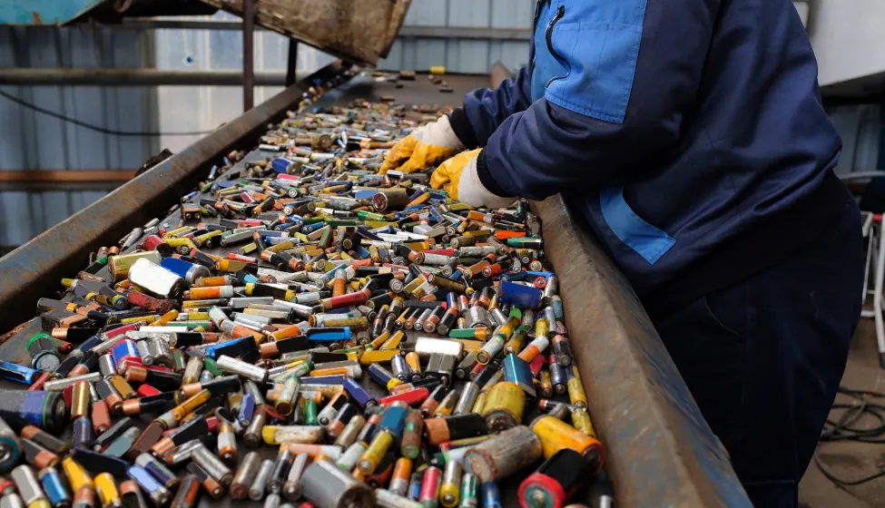 A worker in blue attire and protective gloves sorts through a heap of assorted spent batteries on a conveyor belt at a recycling plant.