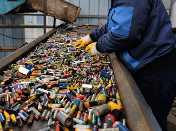 A worker in blue attire and protective gloves sorts through a heap of assorted spent batteries on a conveyor belt at a recycling plant.